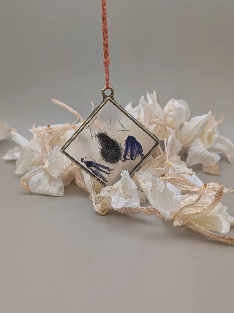 Porte clés mémoriel contenant des poils de blue en forme de larme, dans un cadre losange, en or vieillit avec des fleurs séché violettes